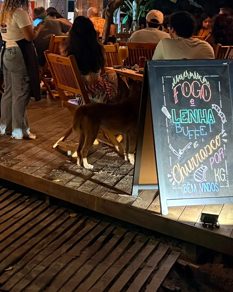 The tables at Fogo e Lenha at night, full of people, with the buffet and churrasco chalkboard in the foreground